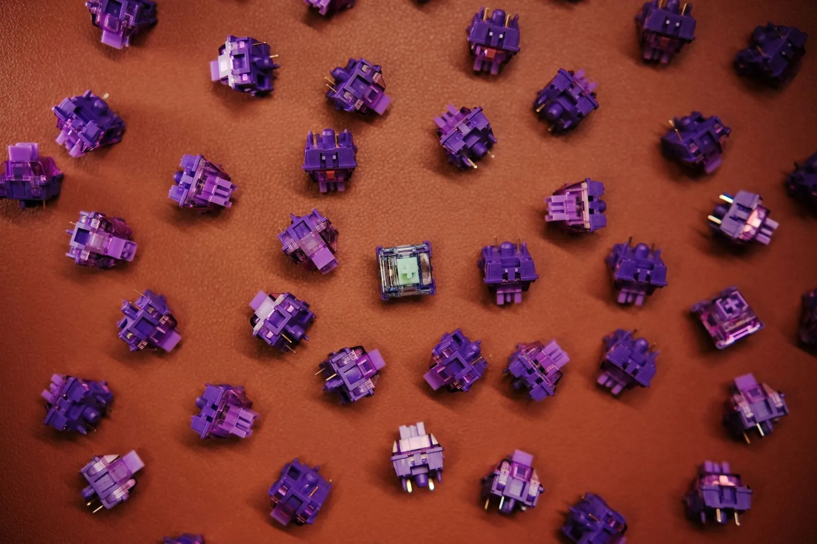 Dozens of loose purple mechanical keyboard switches scattered on brown leather, with one clear-housed switch with a mint-green cross-shaped stem sitting in the middle.