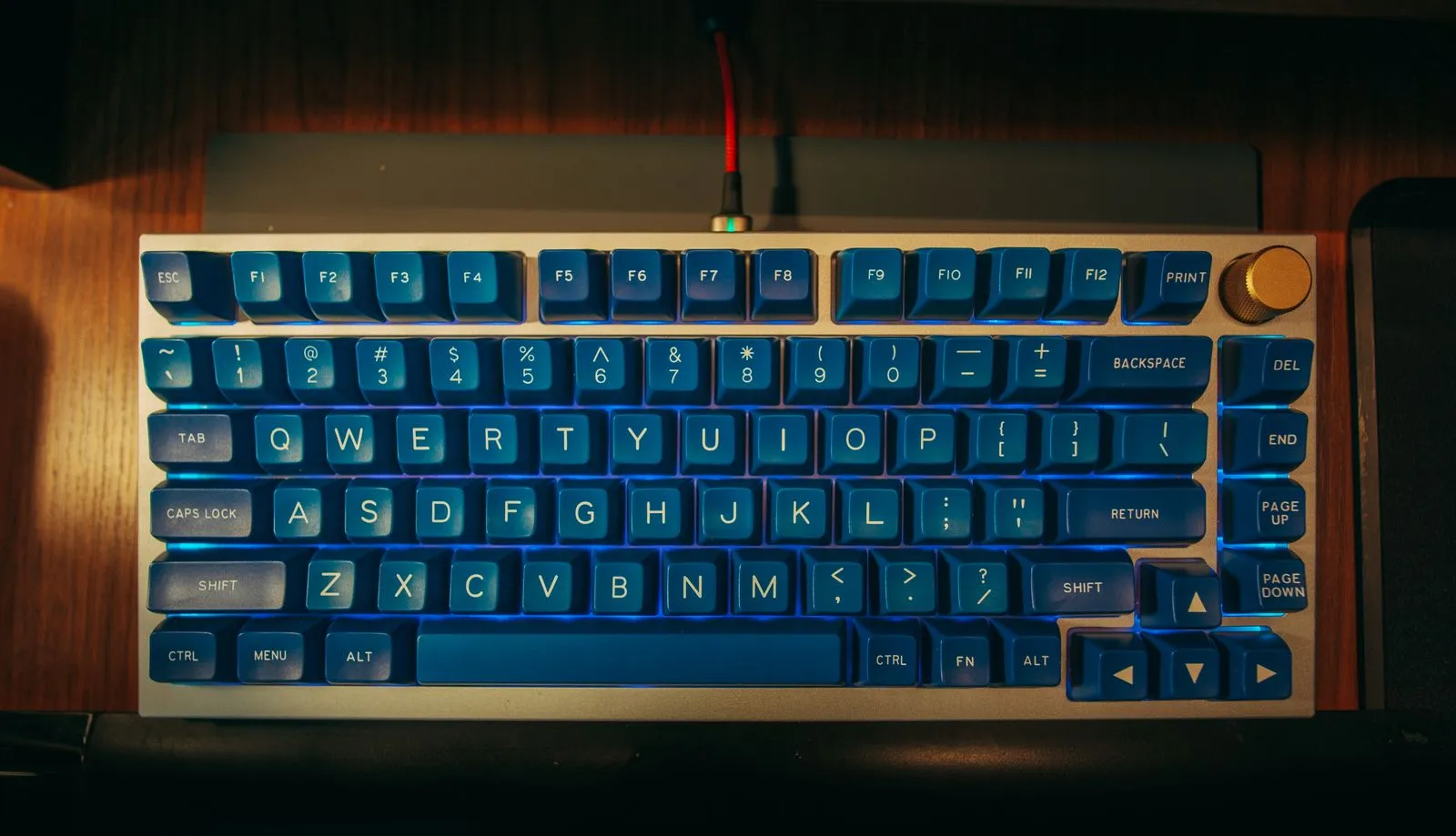 Top-down view of a 75% mechanical keyboard with blue keycaps and a blue underglow in a silver aluminum case, with a gold volume knob and a red coiled cable, on a wooden desk.