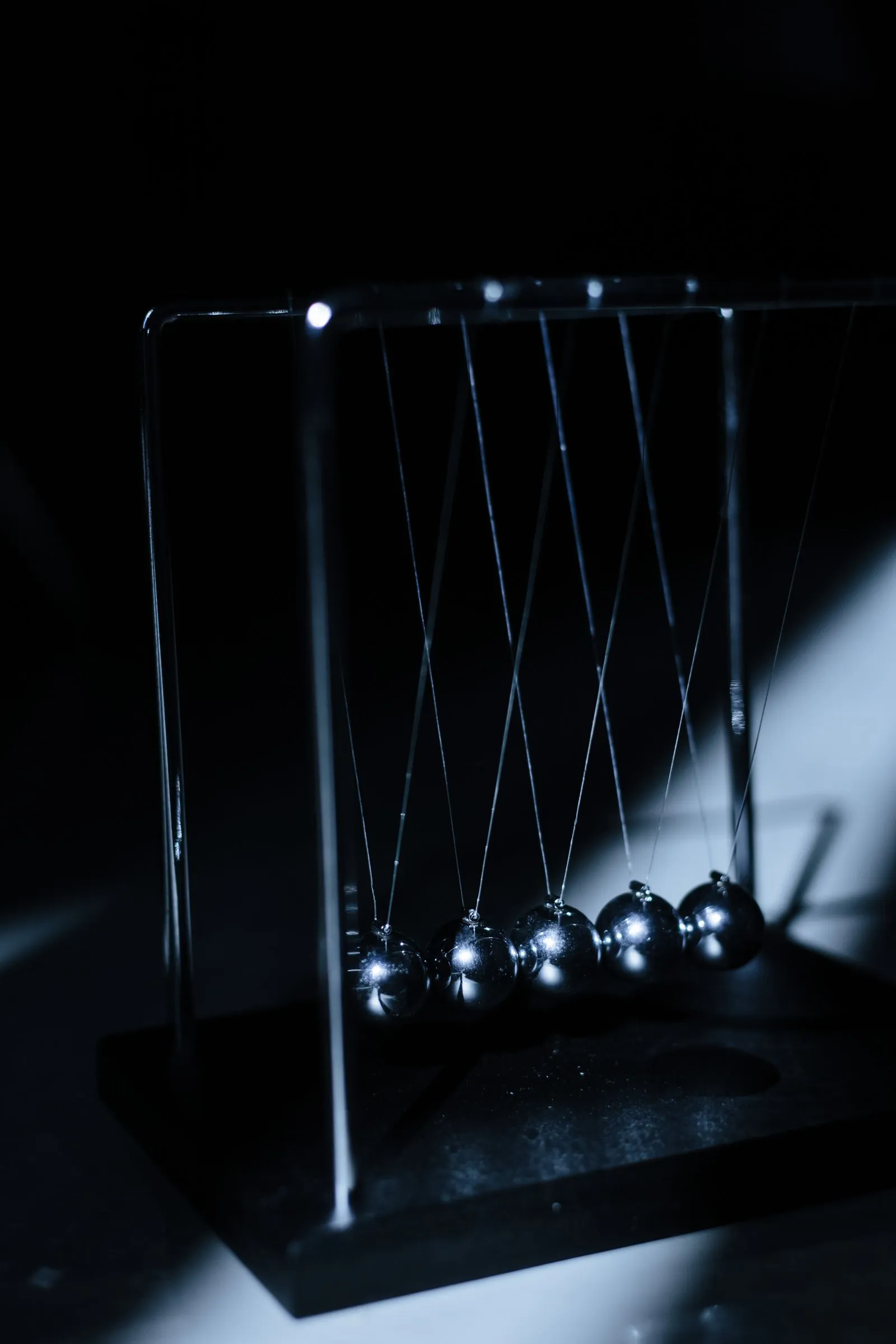 A steel Newton's cradle in hard side light, five polished balls hanging in a row from V-shaped strings over a dark base.